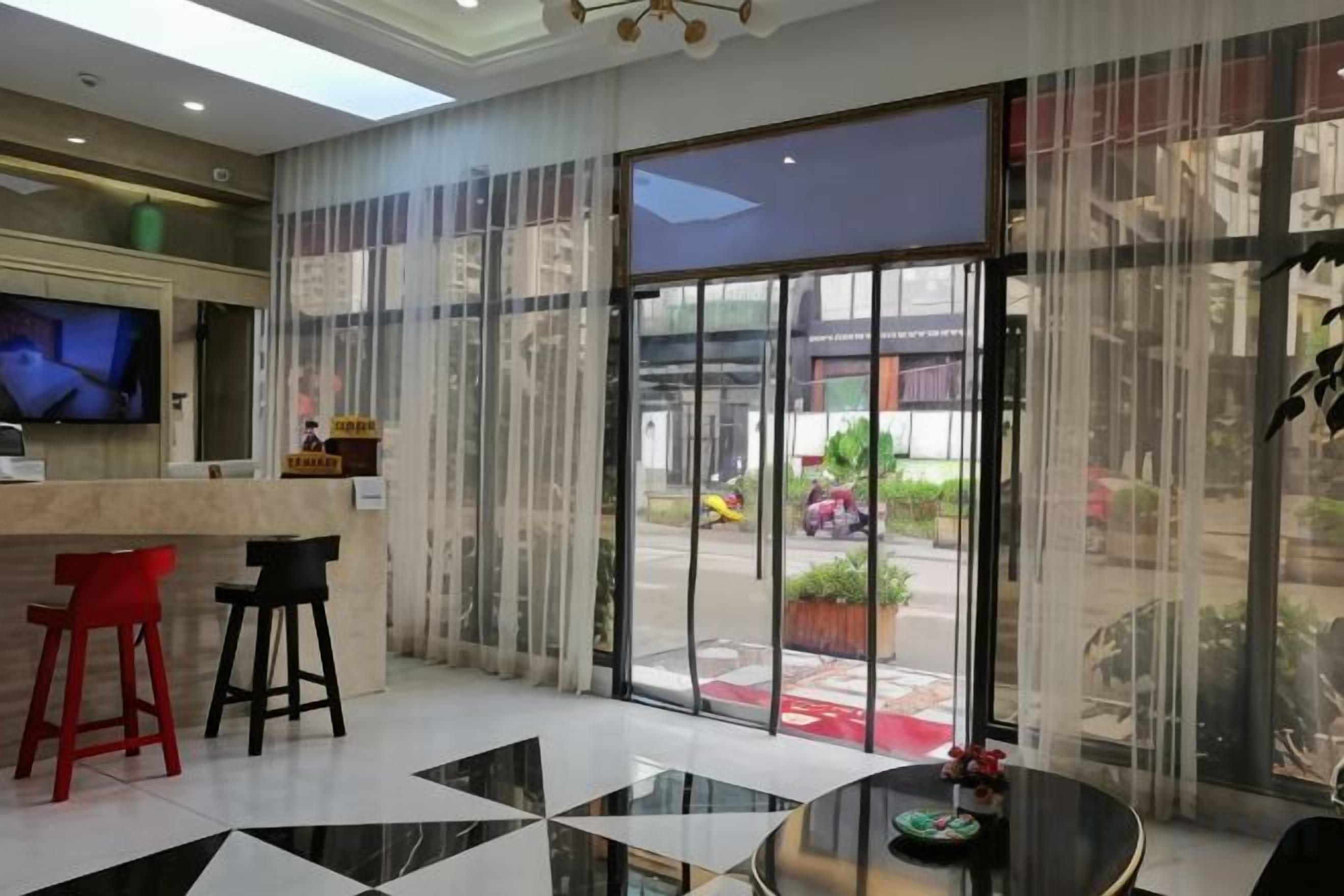A hotel lobby with a reception desk, two bar stools, a round table, and a large glass door leading outside.
