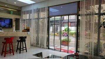 A hotel lobby with a reception desk, two bar stools, a round table, and a large glass door leading outside.