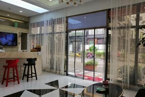 Un hall d’hôtel comprenant un comptoir d’accueil, deux tabourets de bar, une table ronde et une grande porte vitrée donnant sur l’extérieur.