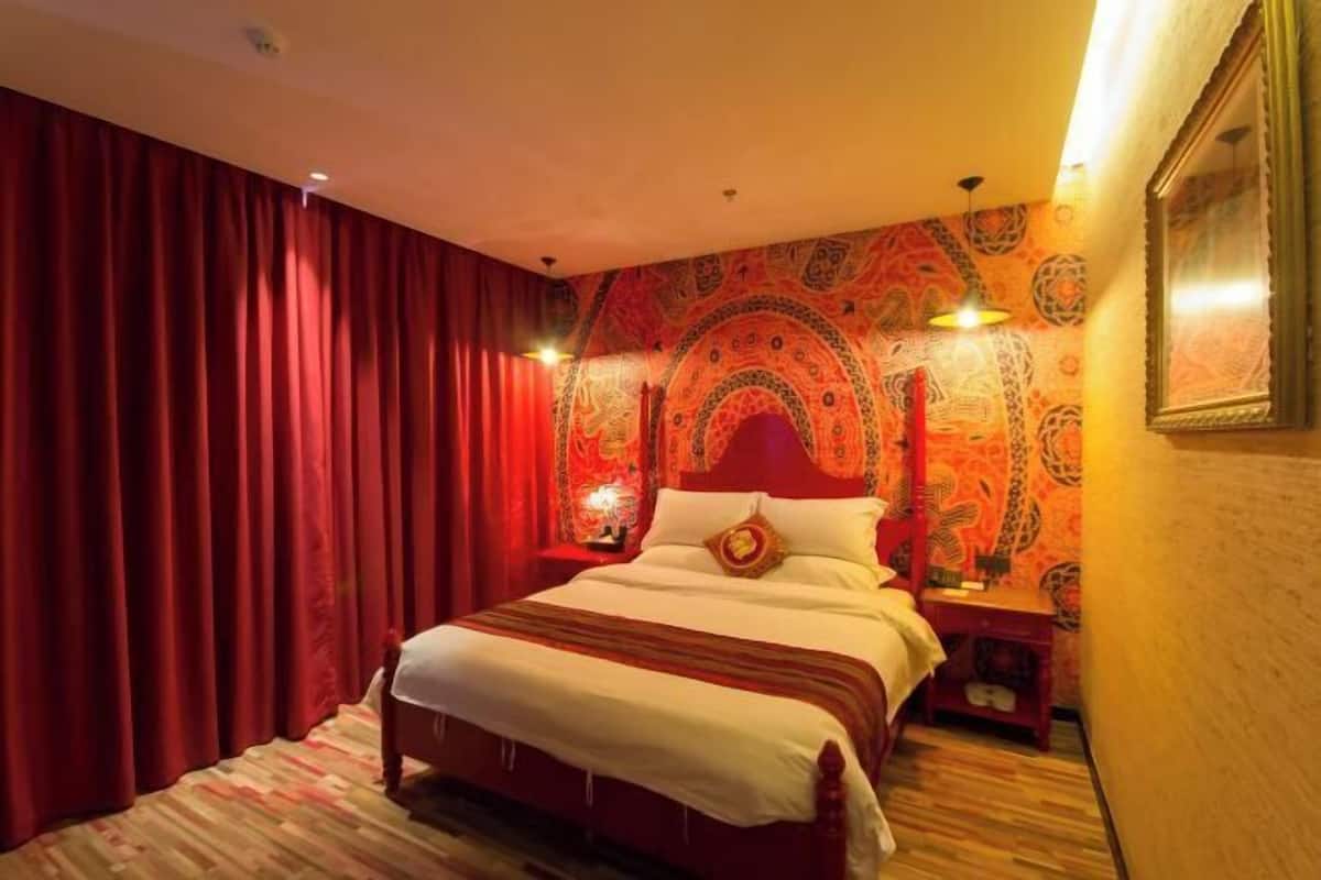 A hotel room with a large bed, red curtains, patterned wallpaper, and a nightstand with a lamp.