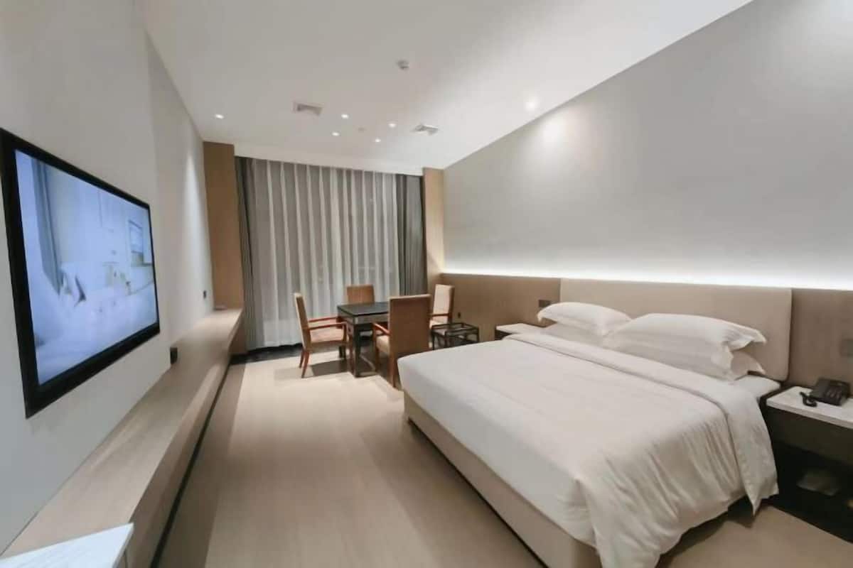 A hotel room with a bed, a desk, a chair, a TV mounted on the wall, and a window with curtains.