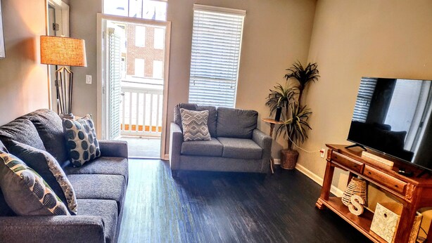 Una sala de estar con piso de madera oscura, un sofá gris, un sillón gris, un televisor sobre un mueble de madera y una planta en maceta.