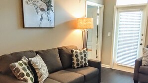 Un salón con piso de madera oscura, un sofá gris con cojines estampados, una lámpara de pie y un cuadro en la pared.
