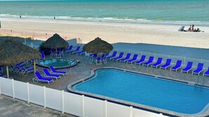 Zona de piscina con sillas reclinables azules y sombrillas de paja, con vista a una playa y una vista despejada del océano.