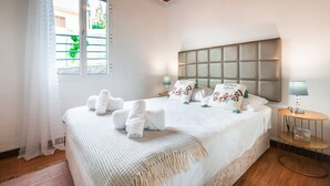 Una cama bien hecha con sábanas blancas y un cabecero decorativo, una mesita de noche con lámpara y una ventana con vistas a zonas verdes.
