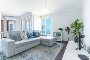 A modern living room with a grey sectional sofa, a patterned rug, a bookshelf, and a view of the sea through large windows. - Sunny Side by Atlantic Holiday (Madalena do Mar)