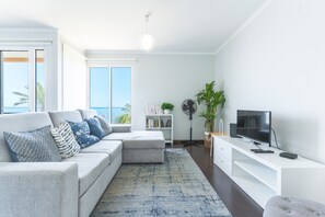 Una sala de estar moderna con un sofá seccional gris, un televisor de pantalla plana y un amplio ventanal con vista al océano.