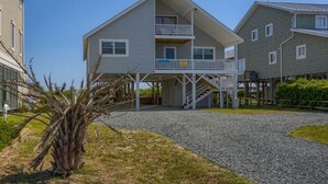A two-story building with a balcony on the upper floor, a staircase leading to the upper level, and a gravel driveway. - A Beach Haven - Oceanfront Home w/Private Pool and Offers Early Check-In/Late Check-Out! (Holden Beach)