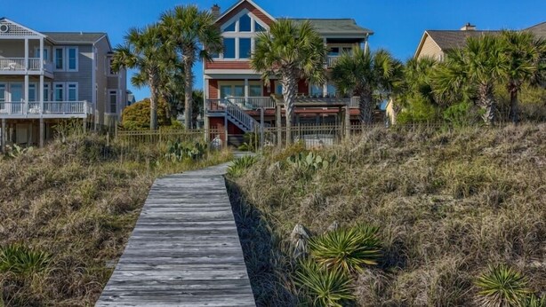 A coastal property with a wooden walkway leading to a house with a balcony, surrounded by palm trees and dry grass.
