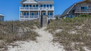 Une maison située en bord de mer, avec un escalier menant à un balcon, entourée de sable et de végétation.
