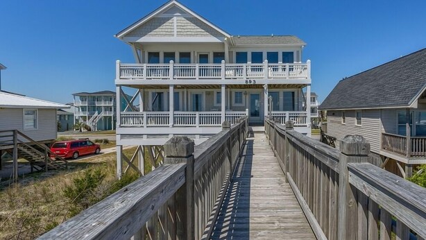 Une maison à deux étages située en bord de mer, avec une terrasse en bois menant à l’entrée.