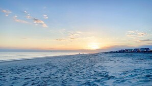 A beach with footprints leading to the water, a sunset in the background, and houses along the shore.