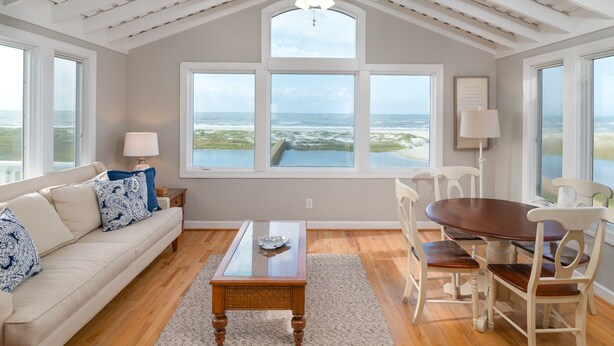 Una habitación amplia con techo alto, ventanas grandes que ofrecen vistas al océano, una mesa de comedor de madera y un sofá confortable.