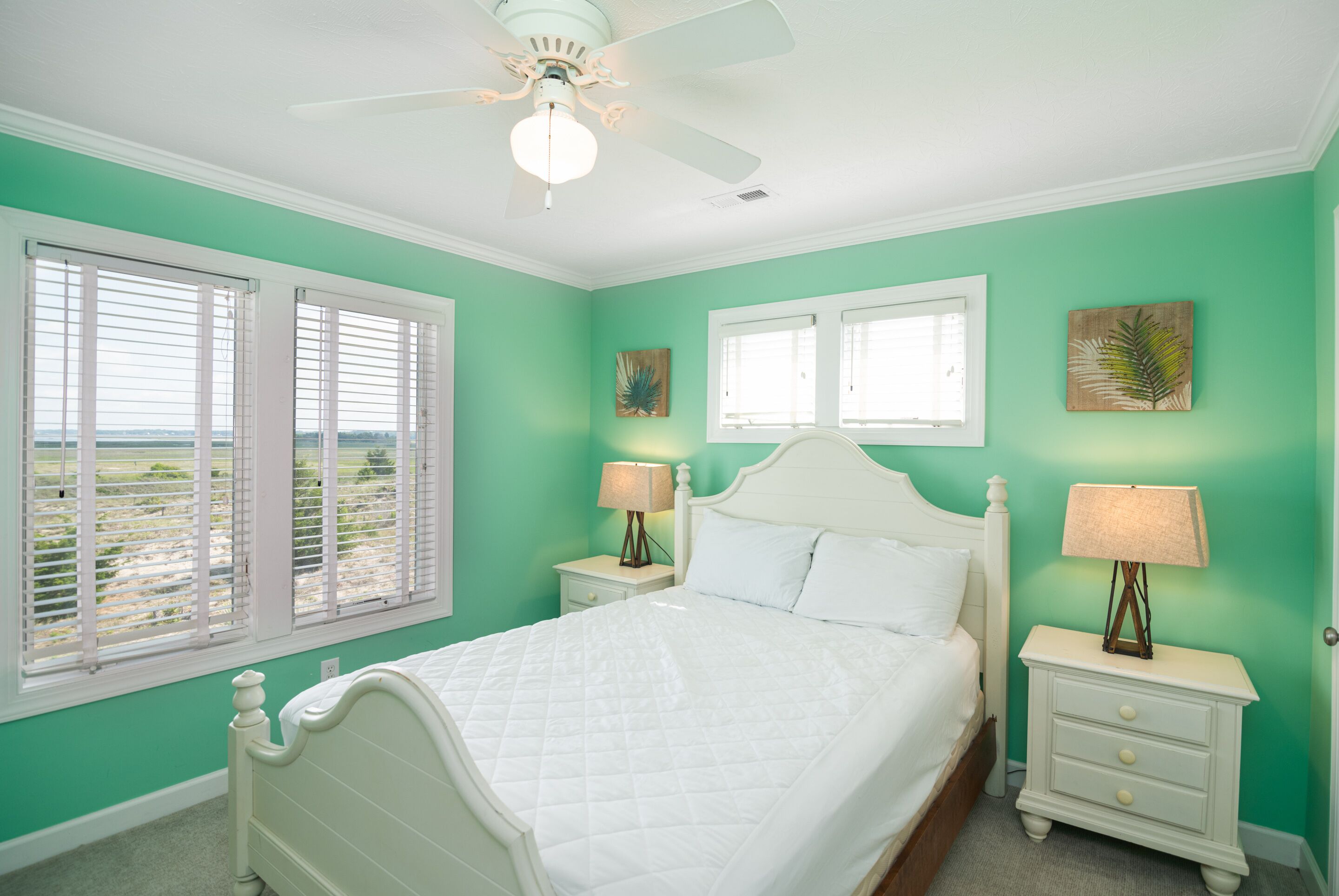 A bedroom with a bed, two bedside tables with lamps, a ceiling fan, and two windows with blinds.