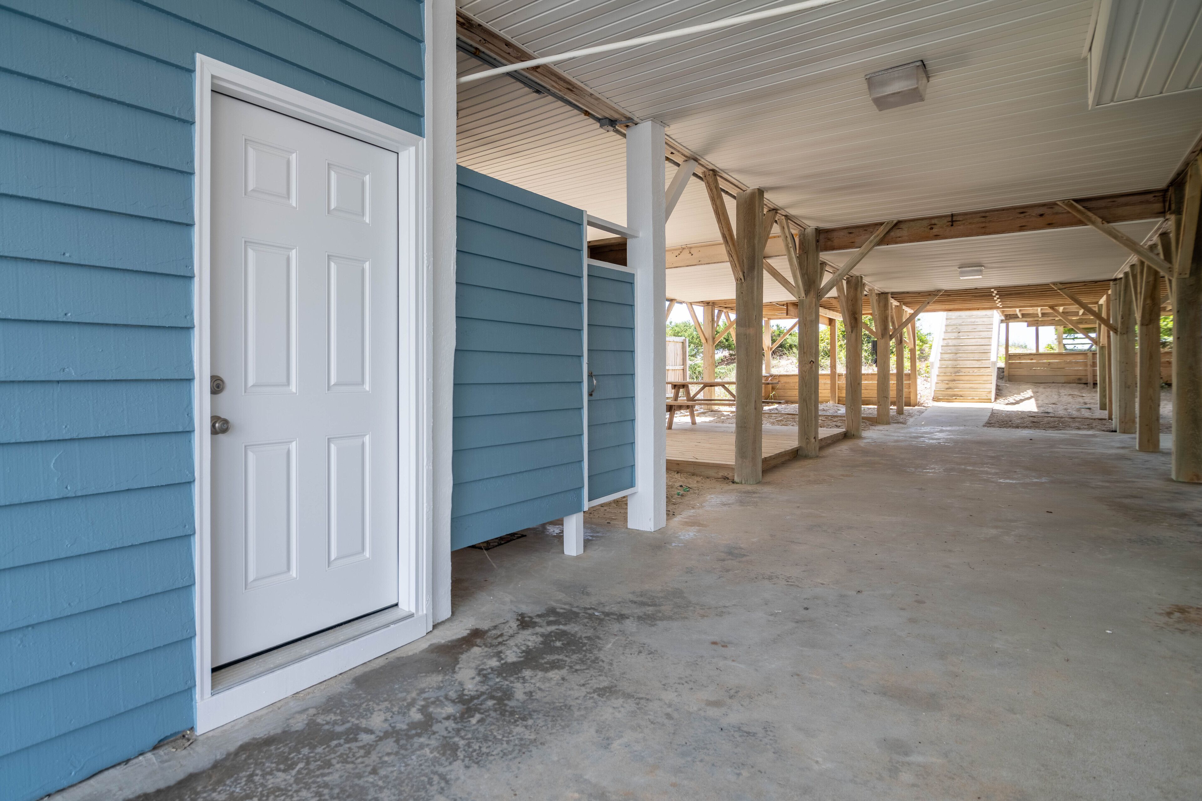 A building with a blue exterior and a white door, featuring a covered outdoor area with wooden beams and a concrete floor.