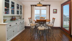 A dining area with a wooden table and four chairs, a white cabinet with glass doors, and a window with a view of the sea.