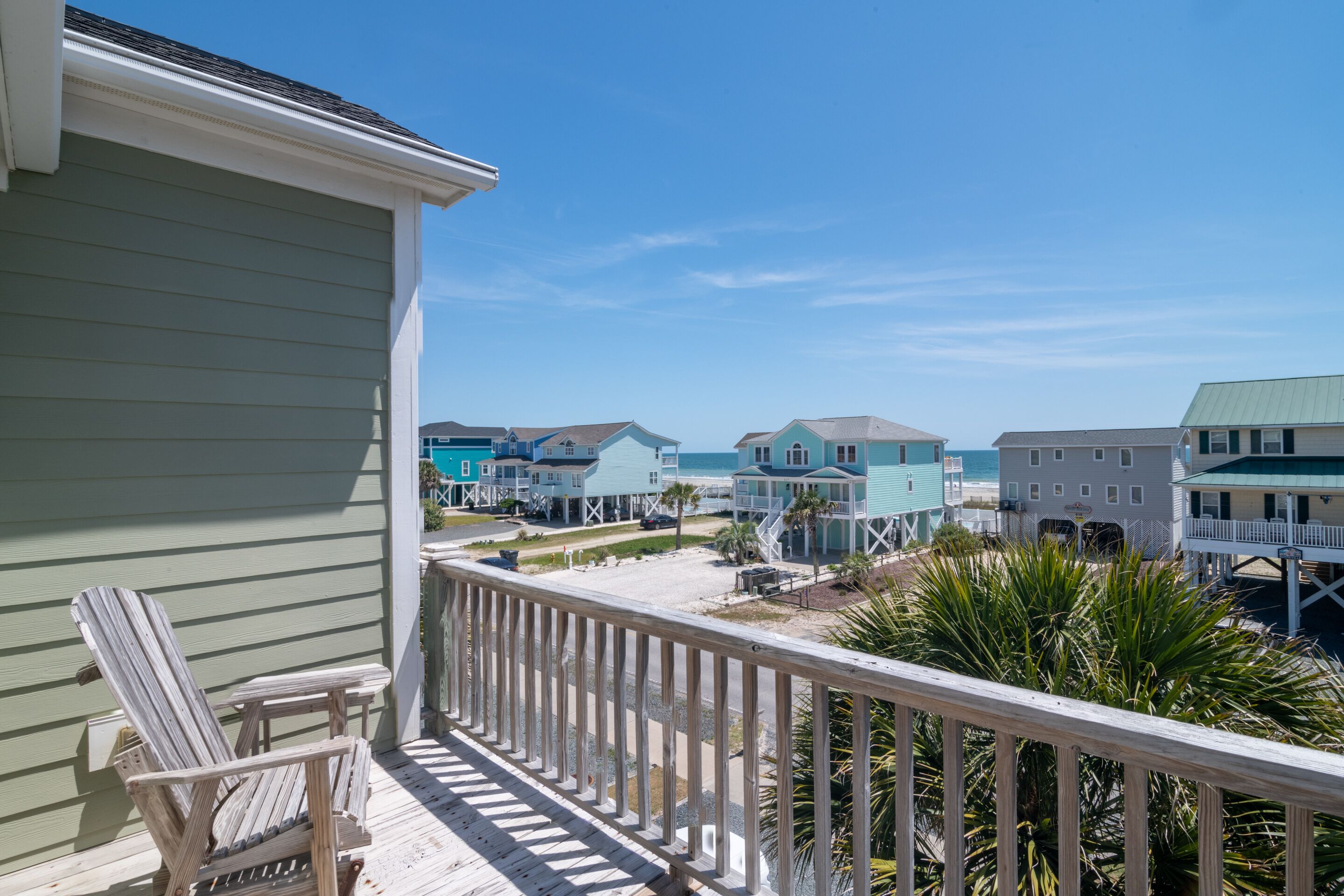 A wooden deck with two Adirondack chairs, a view of beach houses, and a clear blue sky.