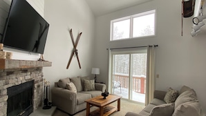 A living room with a fireplace, a mounted deer head, a ceiling fan, a TV, and a large window. - Modern Retreat with Stunning Views. Holiday Valley across the Street! Sleeps 8 (Ellicottville)