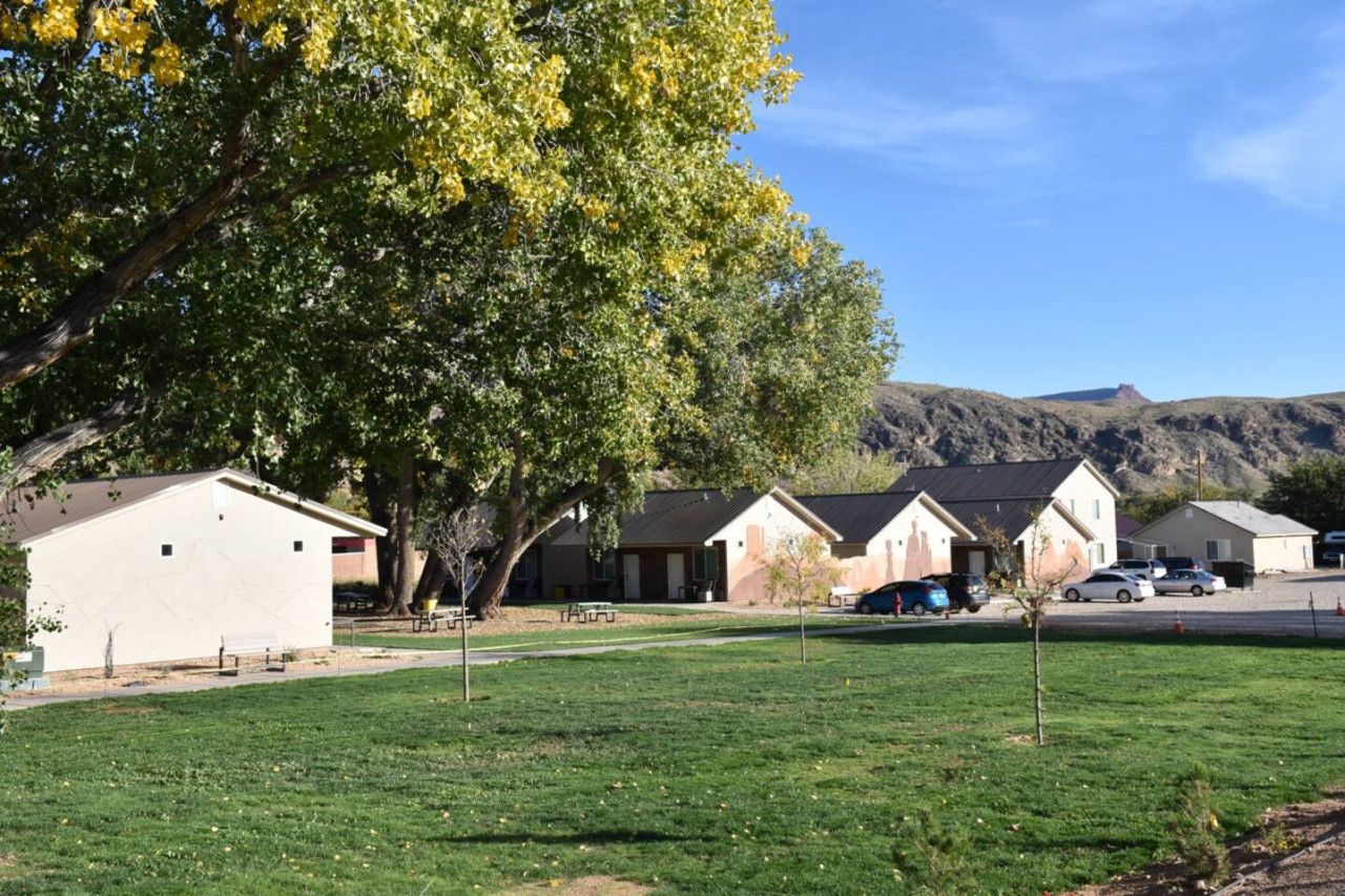 A row of single-story buildings with a grassy area in front, a parking lot, and a mountain in the background.
