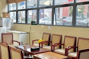 A restaurant interior with a long table and chairs, a window with a view of parked cars, and a small refrigerator. - Pu'er Guopei Hotel (Pu'er)