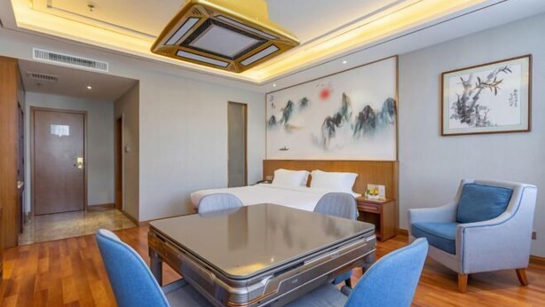 Quarto de hotel com cama, escrivaninha e cadeiras. Há um quadro na parede e uma porta que dá para outro cômodo.