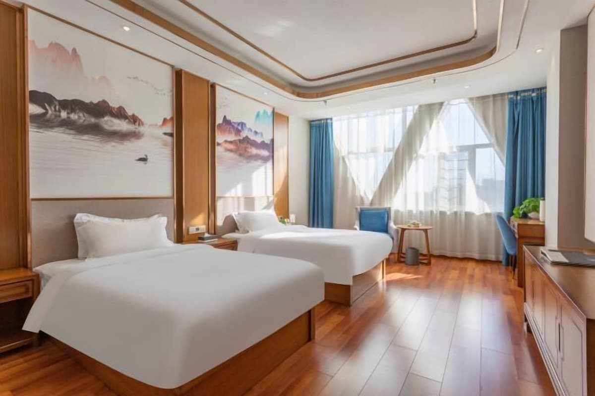 Hotellihuone, jossa on kaksi sänkyä, puulattia, työpöytä, tuoli ja taideteoksia seinillä.