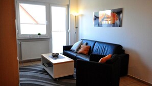 A living room with a black leather sofa, a white coffee table, and a striped rug. There is a window with blinds and a radiator below it.