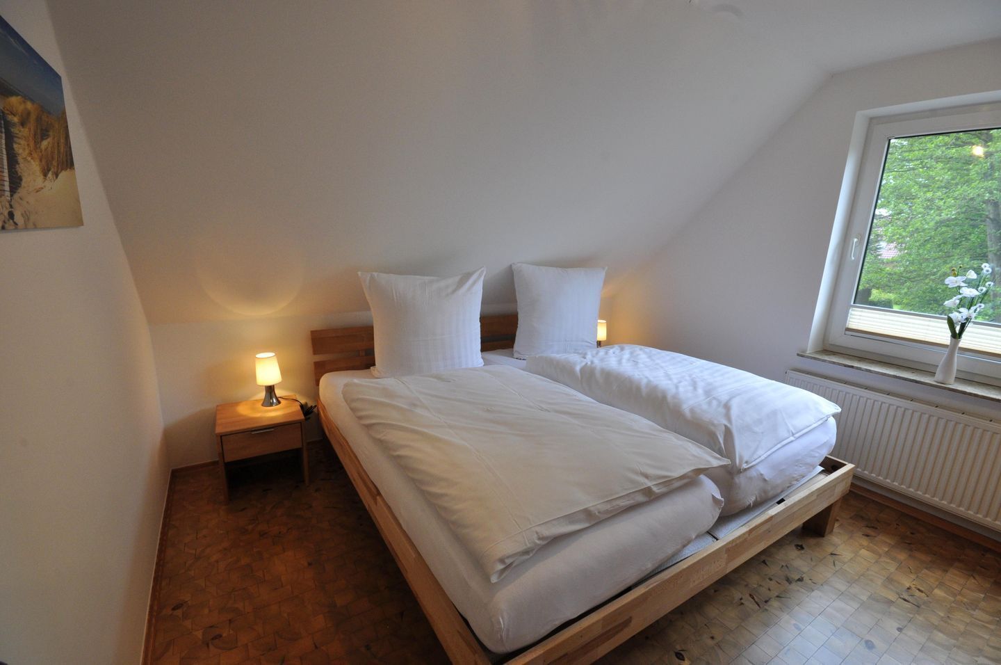 Une chambre avec un lit en bois, une literie blanche, une table de chevet avec une lampe et une fenêtre donnant sur un paysage verdoyant.