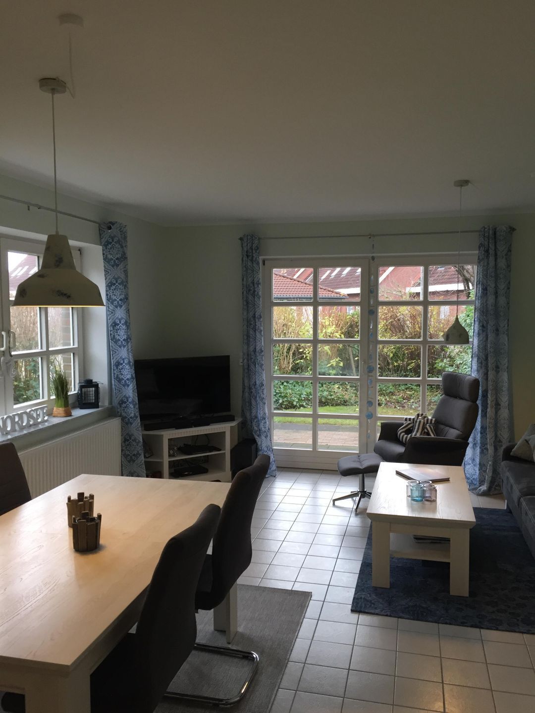 Sala de estar com televisão, mesa de jantar, cadeiras, sofá e mesa de centro. Há uma janela grande com cortinas e uma porta que dá para o exterior.