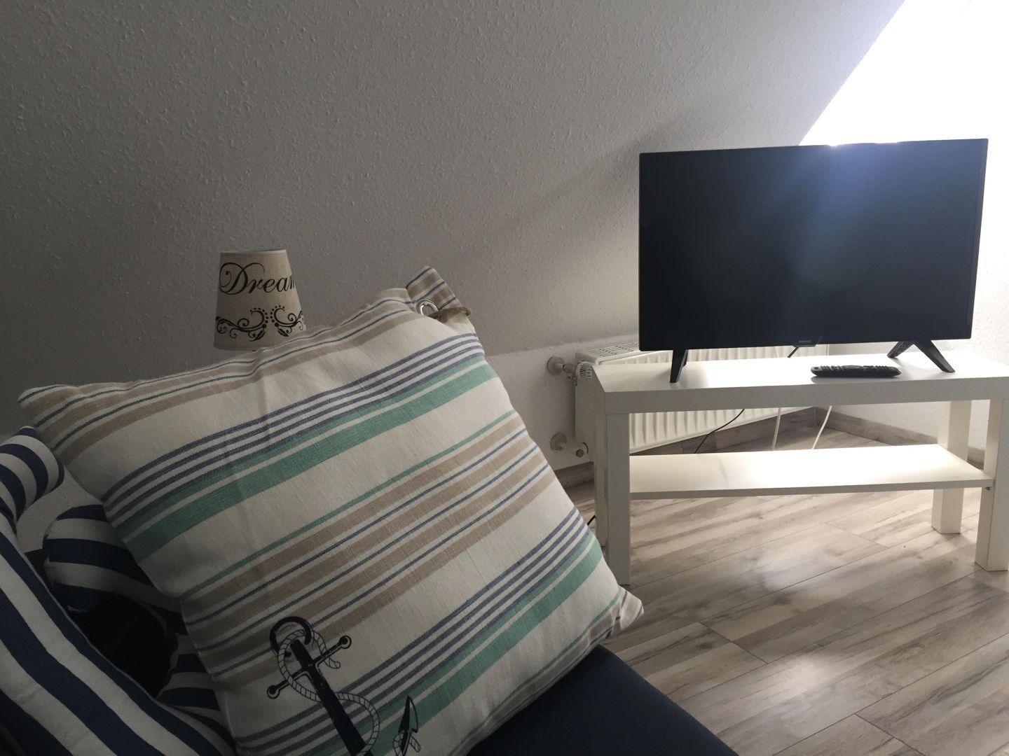 Una habitación con un cojín a rayas, una mesita blanca pequeña con un televisor y un control remoto, y un radiador.