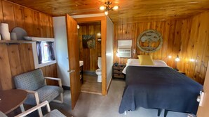 Una habitación de cabaña de madera con cama, una silla, una mesita, un ventanal con cortinas, aire acondicionado de pared y un adorno en la pared.