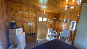 Una habitación con paredes de madera, una cama, un pequeño rincón de cocina, dos sillas y un ventilador de techo.