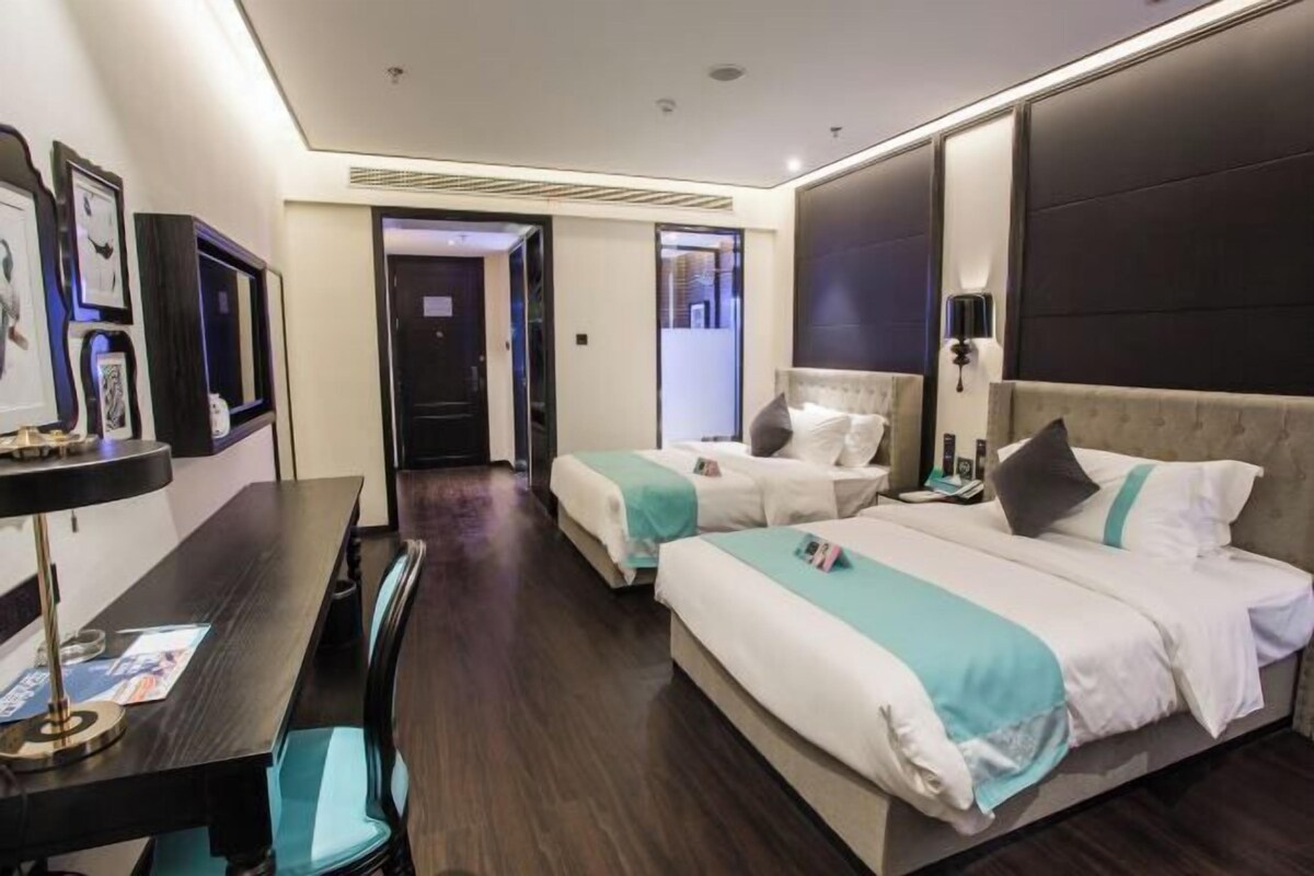 A hotel room with two beds, a desk, and a chair. There is a door leading to another room and a wall with framed pictures.