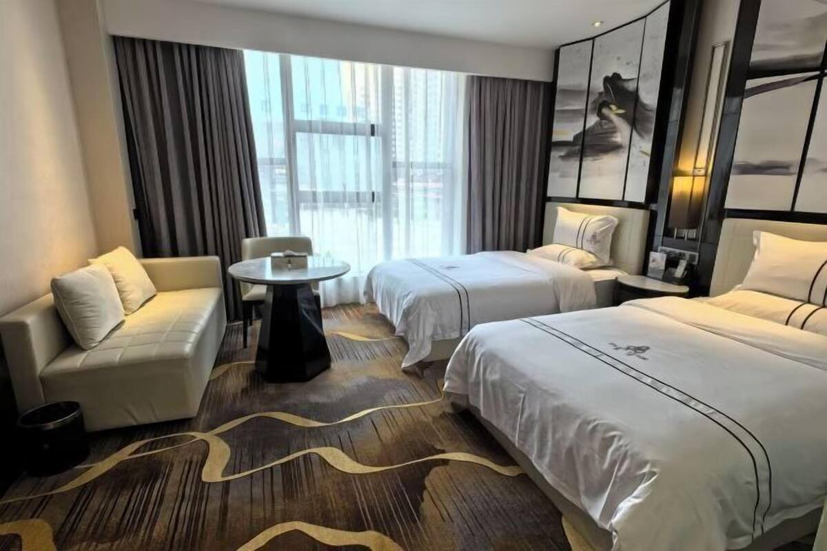 Hotellihuoneessa on sohva, kaksi sänkyä, pieni pöytä ja suuri ikkuna, jossa on verhot.
