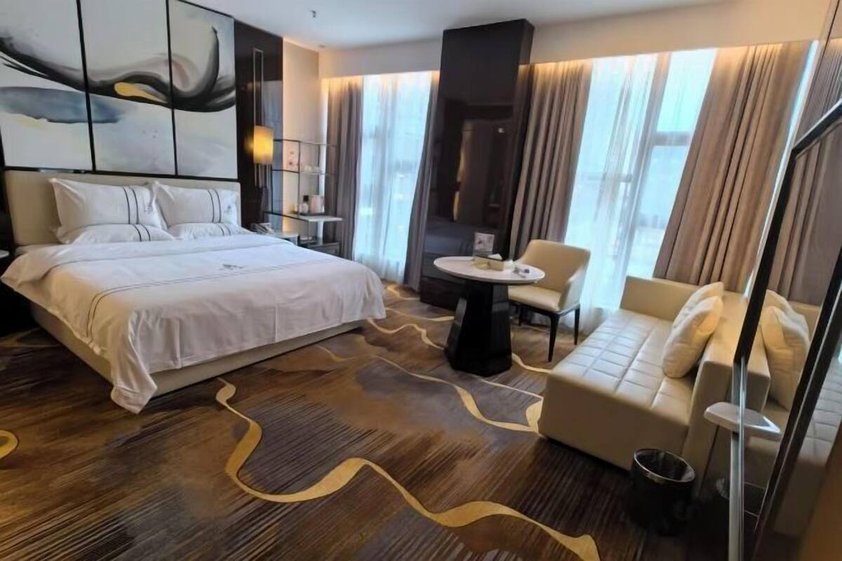 Moderni hotellihuone, jossa on suuri sänky, sohva, pieni pöytä ja suuret ikkunat, joista avautuu näkymä kaupunkiin.