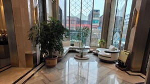 Eine moderne Lobby mit poliertem Marmorboden, einer großen Kübelpflanze, einer Sitzecke mit einem weißen Sofa und Blick auf die Stadt durch große Fenster.