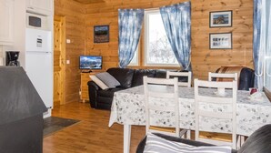 Ein gemütliches Blockhaus-Interieur mit Holzvertäfelung, einem Esstisch mit Stühlen, einer schwarzen Ledercouch, einem Fernseher und einer Küchenzeile mit Kühlschrank und Mikrowelle.