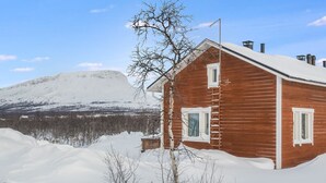 Eine Holzhütte mit verschneitem Dach, einer Leiter und Blick auf einen verschneiten Berg im Hintergrund.