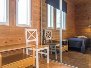 Interno di una baita in legno con una scrivania, due sedie, un letto e una finestra.