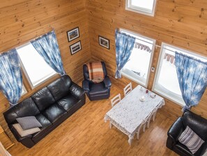 A cozy cabin interior with a leather sofa, a small table, and two chairs. There are two windows with curtains, and the walls are wooden.
