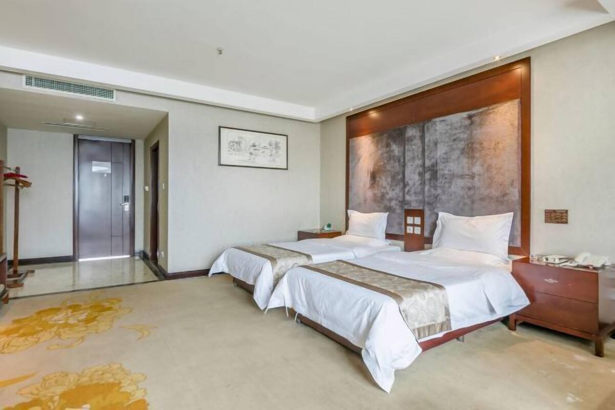 A hotel room with two beds, a wooden headboard, a bedside table with a lamp, and a large abstract painting on the wall.