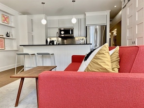 Una cocina moderna con armarios blancos, un refrigerador de acero inoxidable y un microondas. Hay un sofá rojo con una manta estampada y una mesa de madera con dos sillas.