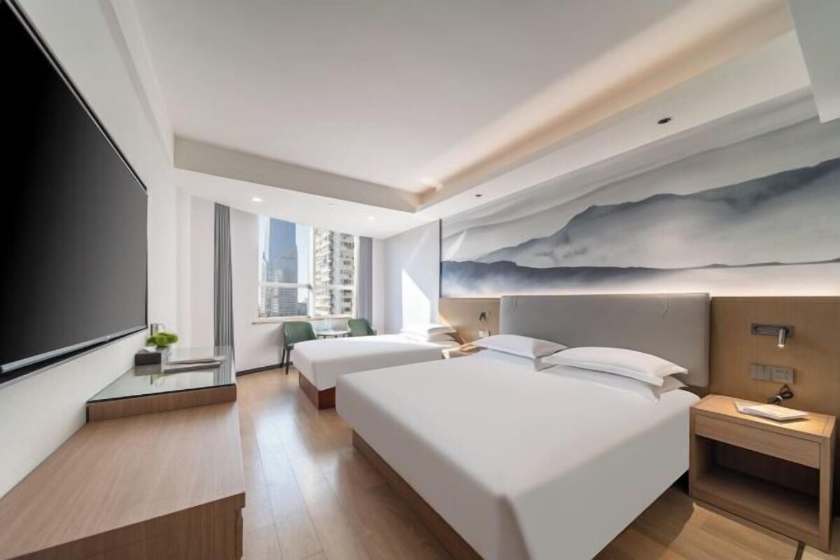 A modern hotel room with two beds, a large TV, a desk with a chair, and a city view.