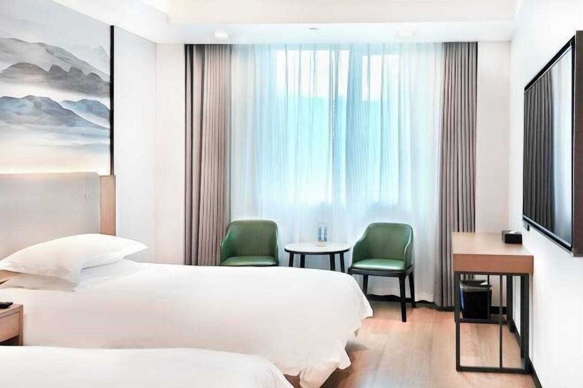 A hotel room with two beds, a desk, two chairs, a TV, and a large window with curtains.