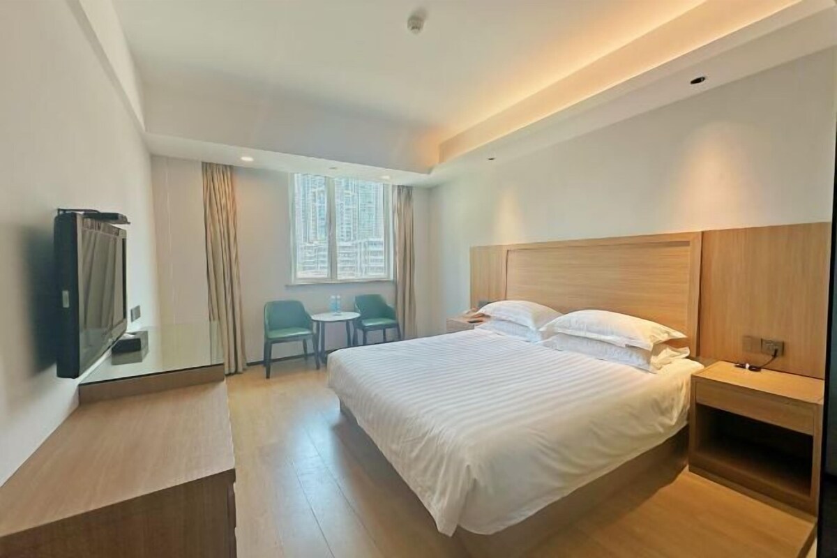 A hotel room with a large bed, a bedside table, a TV, a window with curtains, and a small table with chairs.