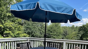 A round table with wicker chairs and a blue umbrella on a wooden deck with a white railing. - Sacandaga Lake Adirondack Escape (Gloversville)