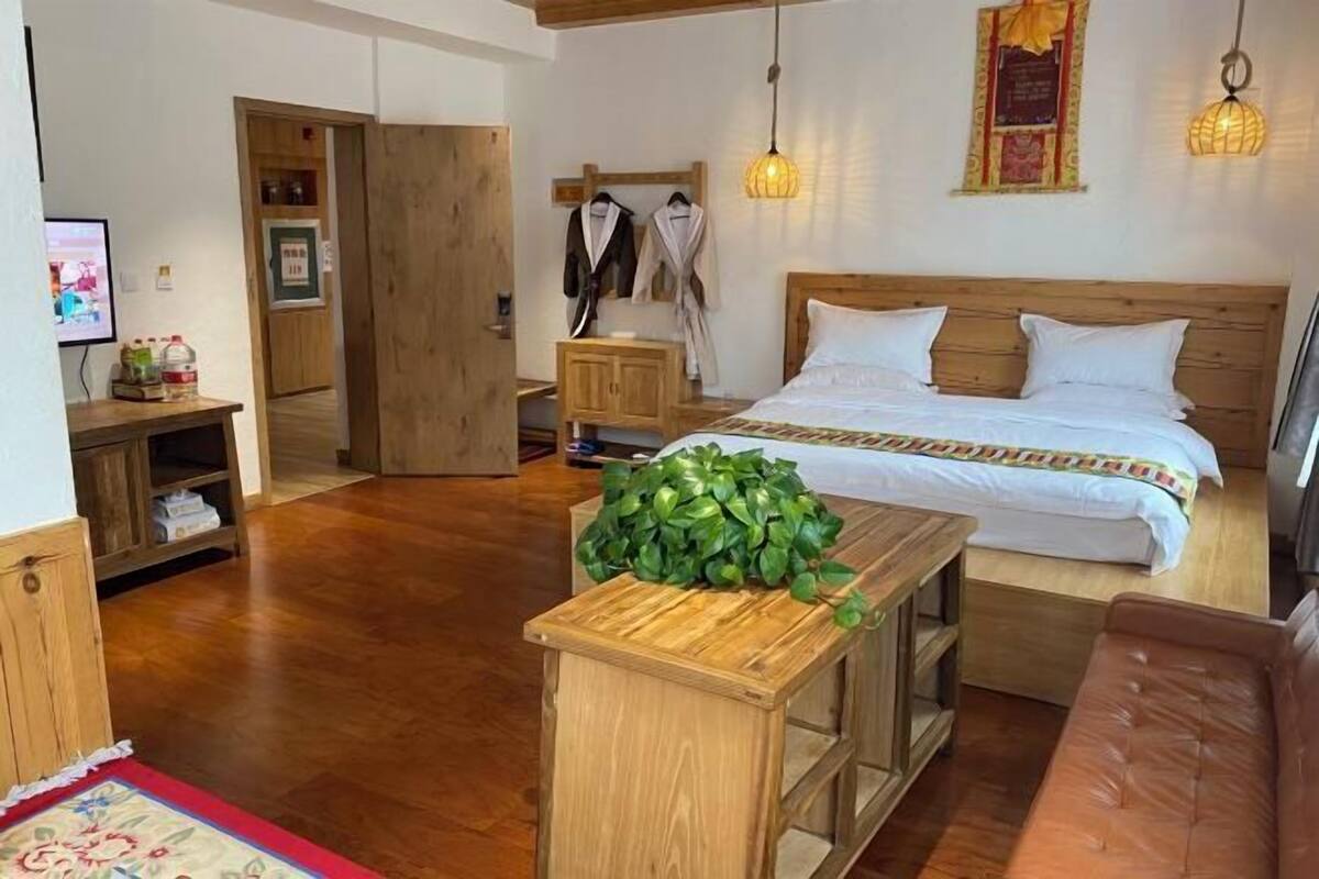 A bedroom with a wooden bed, a bedside table, a small plant, a wooden bench, a red and white patterned rug, and a brown leather couch.