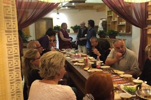 Un grupo de personas sentadas alrededor de una larga mesa de comedor con comida y bebidas, en una habitación con cortinas estampadas y una estantería con plantas.