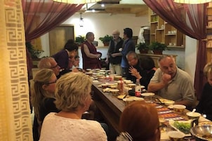 Un grupo de personas sentadas alrededor de una larga mesa de comedor con comida y bebidas, en una habitación con cortinas estampadas y una estantería con plantas.