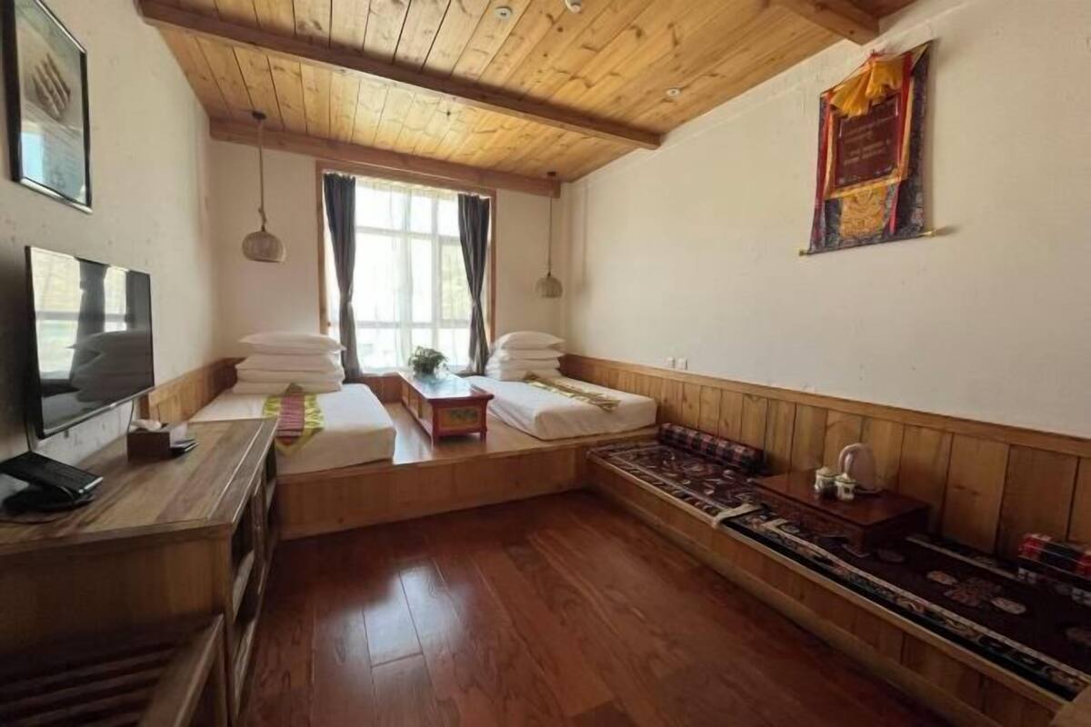 A room with wooden flooring, a bed, a desk with a computer, a window with curtains, and a wall-mounted decoration.
